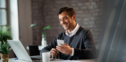 Pessoa sorrindo enquanto usa celular e laptop em mesa de escritório — uso típico do certificado digital e-CPF (imagem oficial Soluti)